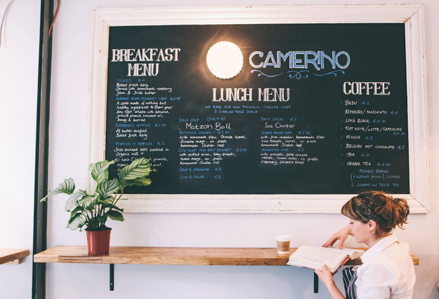 Caryna Camerino sits with a coffee reading in front of a large menu board