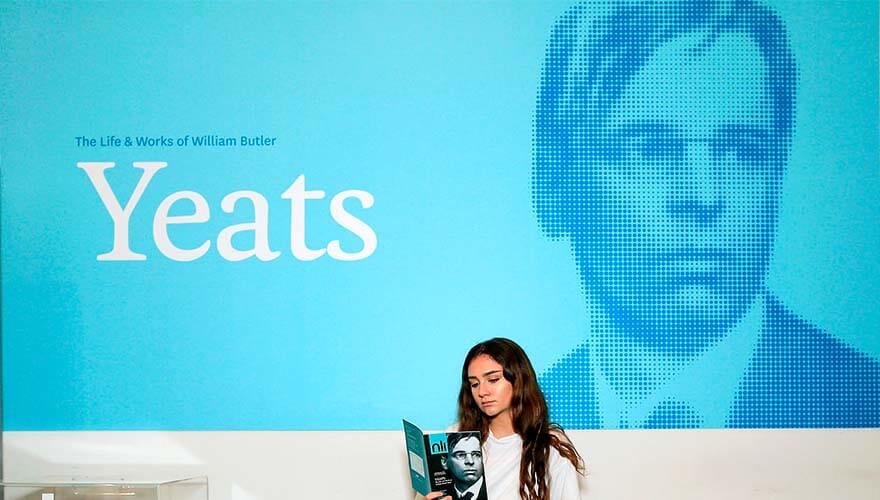 a woman reads a programmes in front of a blue display screen for the WB Yeats exhibition